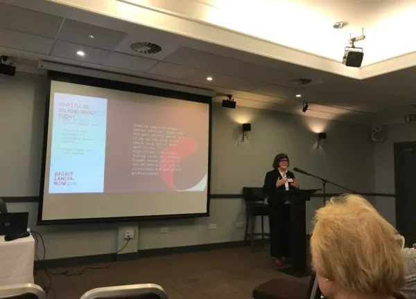 A speaker addressing the conference from a lectern, a slide on the screen beside her.