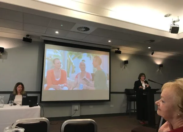 The conference screen showing a photograph of a group of women, with the speaker at the lectern.