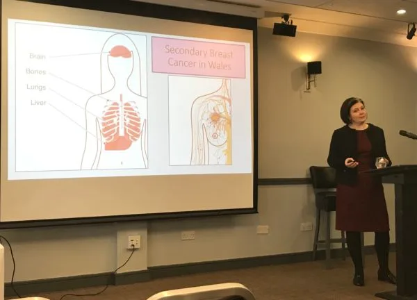 A speaker beside a slide showing where secondary breast cancer spreads in the body: brain, bones, lungs and liver.