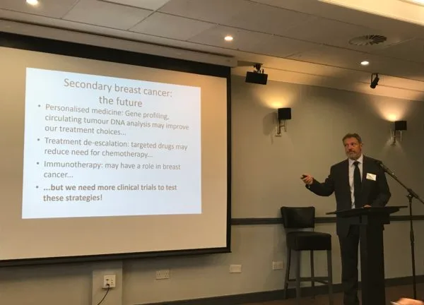 A speaker beside a slide headed Secondary breast cancer: the future, listing personalised medicine, gene profiling, circulating tumour DNA, treatment de-escalation and immunotherapy, and ending: but we need more clinical trials to test these strategies.
