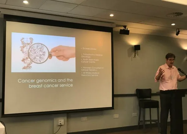 A speaker presenting a slide headed: cancer genomics and the breast cancer service.