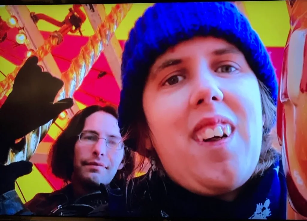 Kit in a bright blue woolly hat, laughing into the camera at a funfair with a friend just behind.