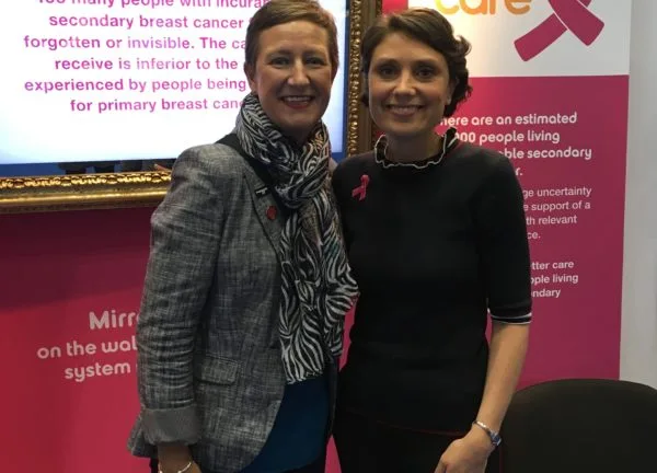 Two campaigners in front of a Breast Cancer Care stand, beside a screen about people with secondary breast cancer being forgotten.