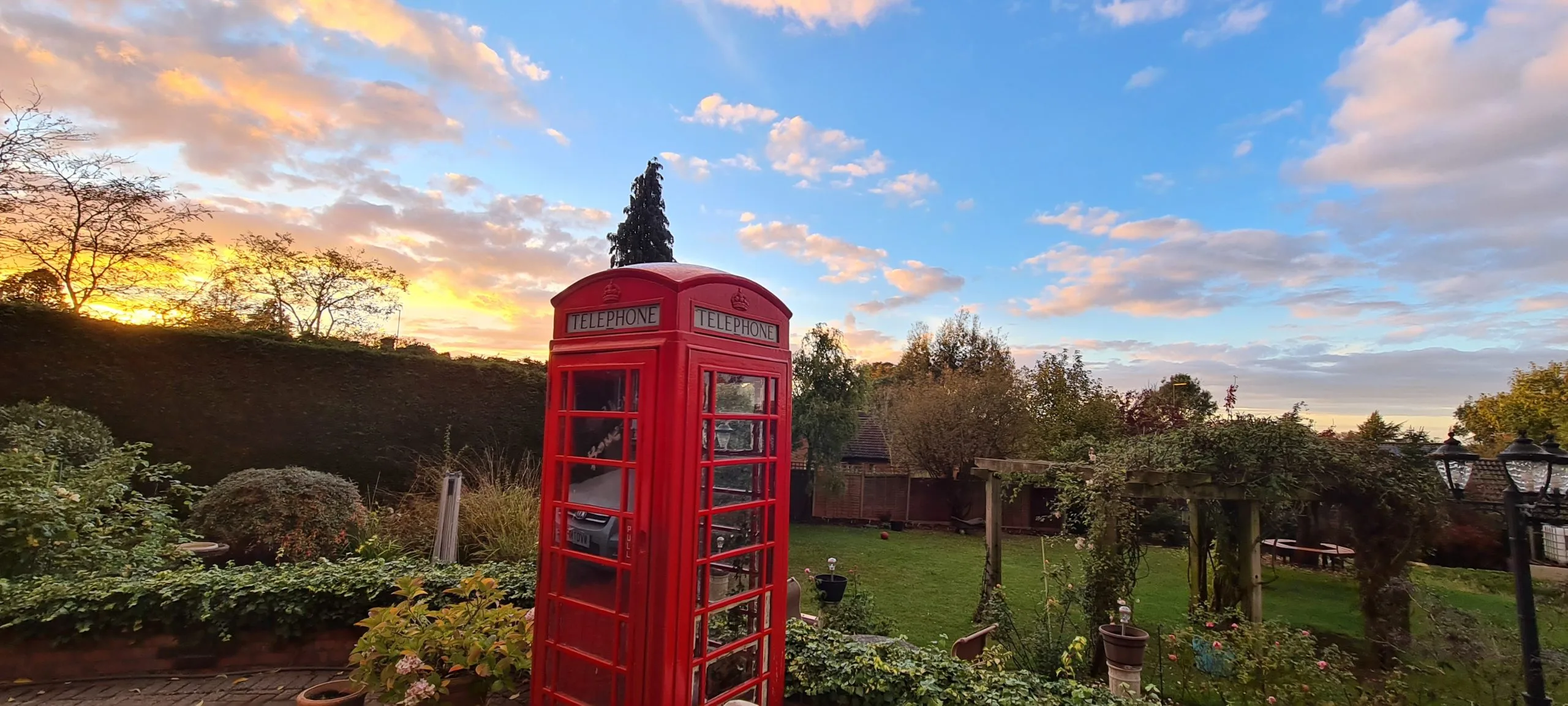 A garden at sunset, a red telephone box standing among the shrubs under a bank of cloud lit orange.