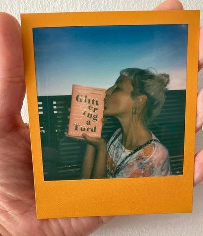 A Polaroid held up in somebody hand, showing a woman kissing a copy of the book Glittering a Turd.