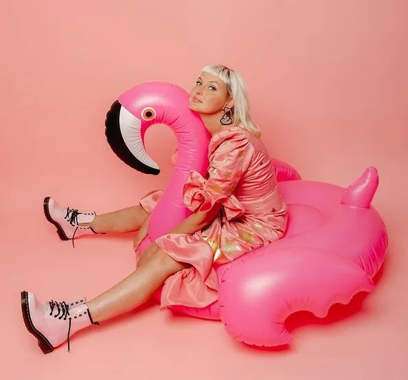 Kris Hallenga lying back across an inflatable flamingo in a pink dress and white boots.