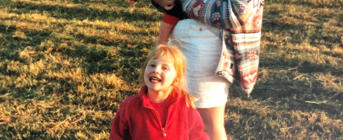 A woman holding a baby in a hay field at golden hour, a small girl in a red coat beside her.