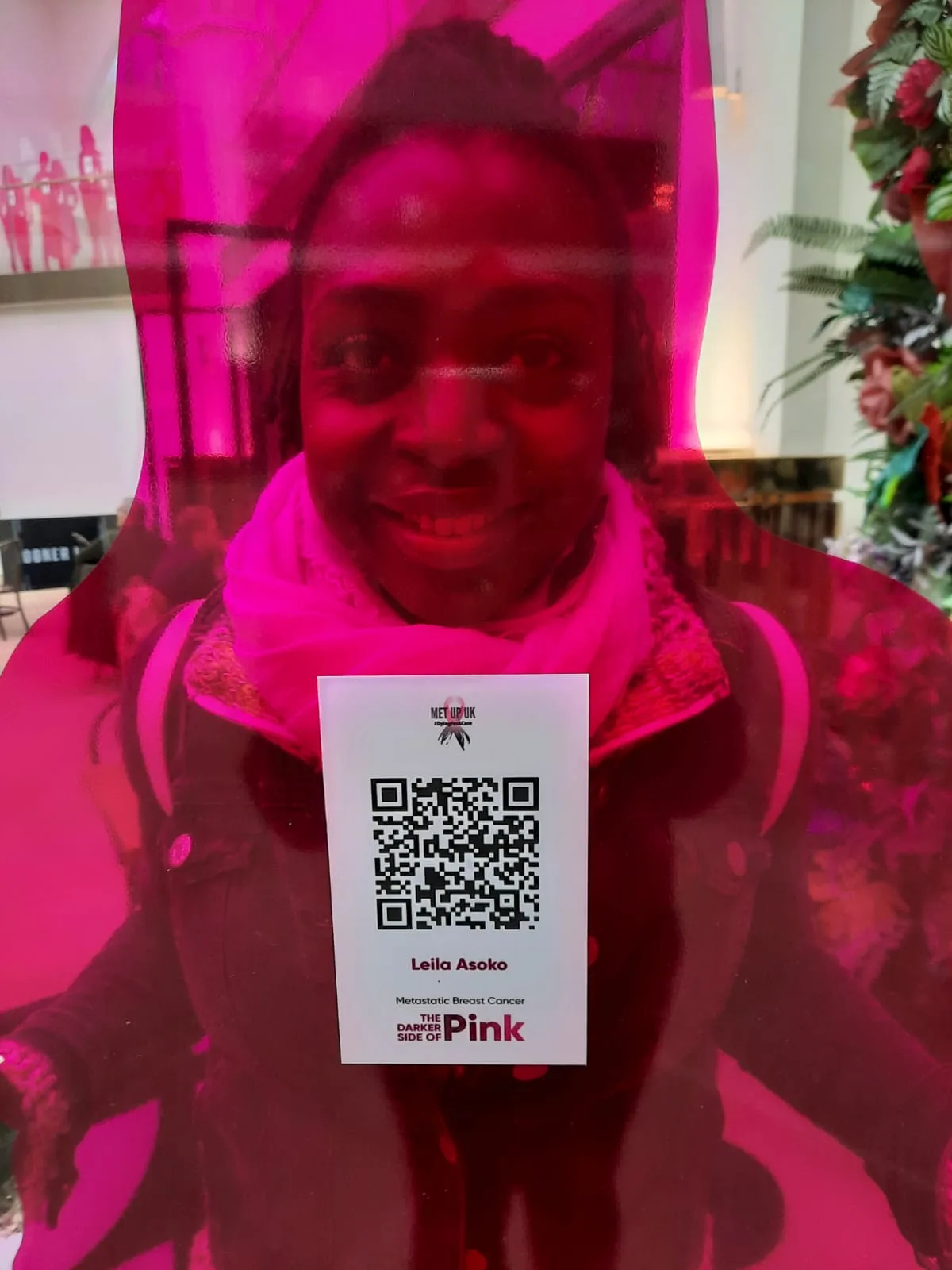 Leila Asoko behind a bright pink Darker Side of Pink display, holding up a card with a QR code that links to her story.