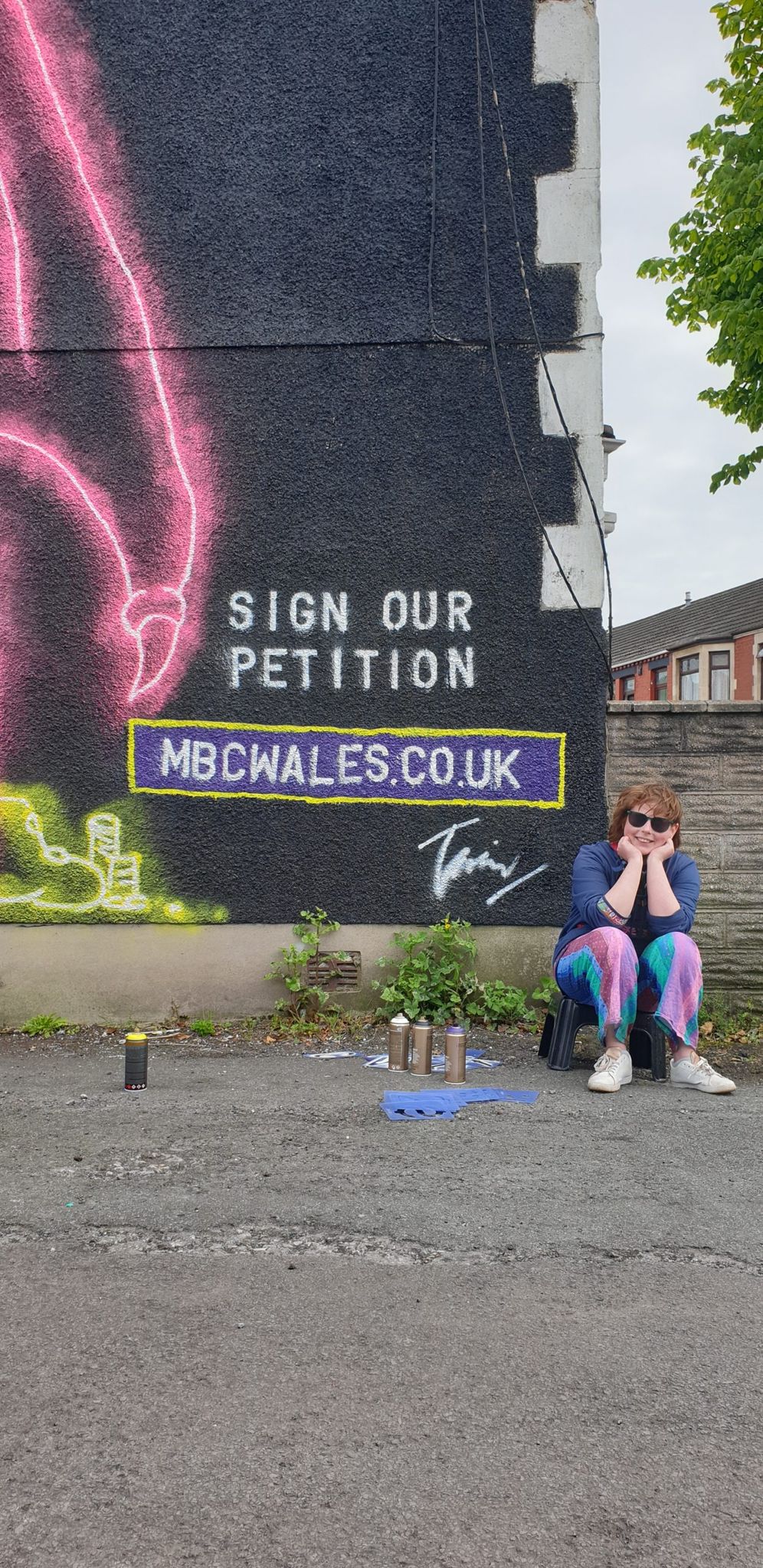 Tassia Haines sitting beneath the mural, beside the painted words Sign our petition, mbcwales.co.uk.