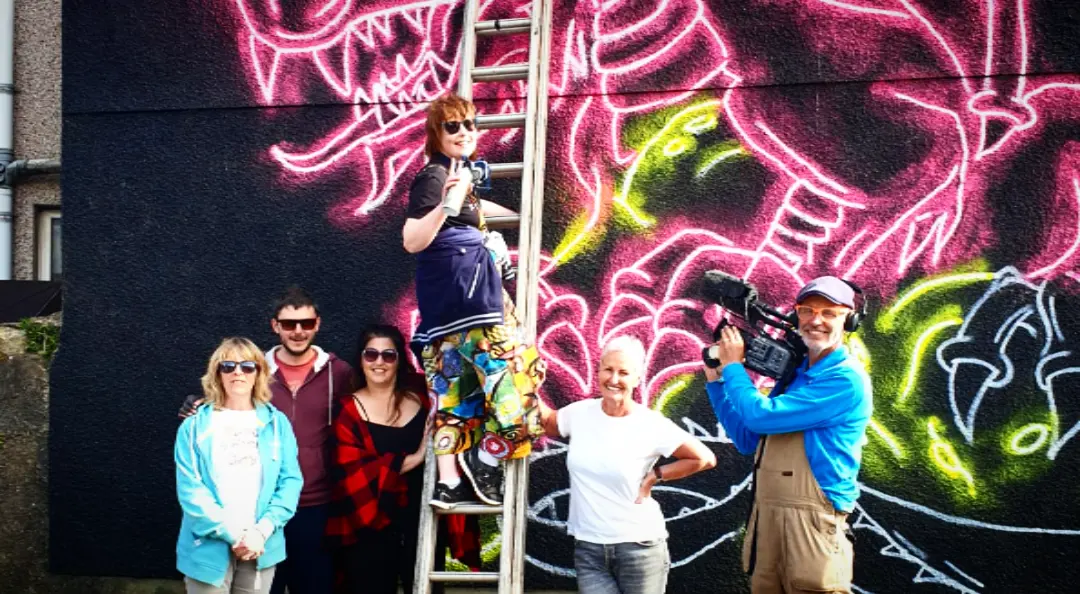 The group who made the mural posed in front of it on a ladder, with a camera operator filming them.