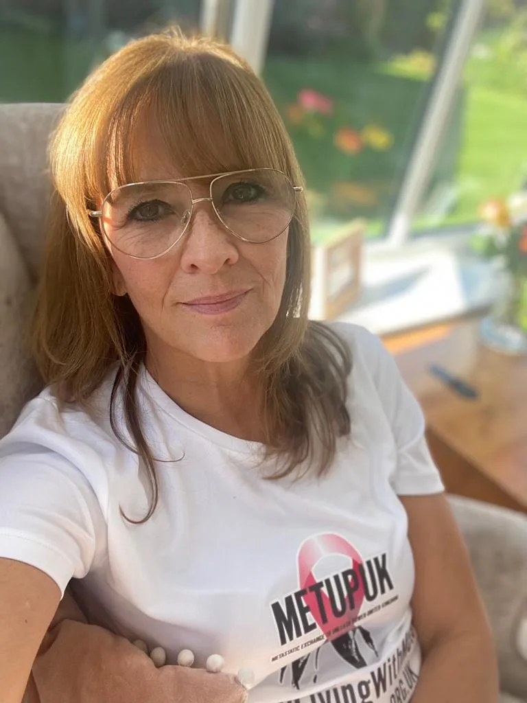 Laura Ashurst in a METUPUK Busy Living With Mets T-shirt, sitting in a conservatory.