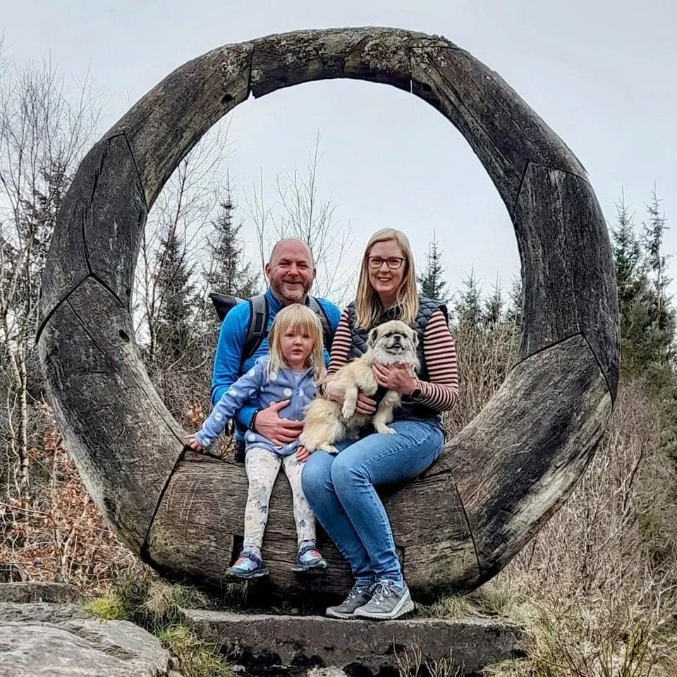 Emma Saville with her family and their dog, sitting inside a large circular wooden sculpture on a hillside.