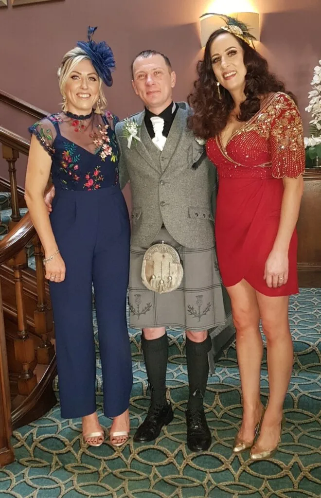Three people dressed for a wedding on a staircase, two in fascinators and one in a kilt.