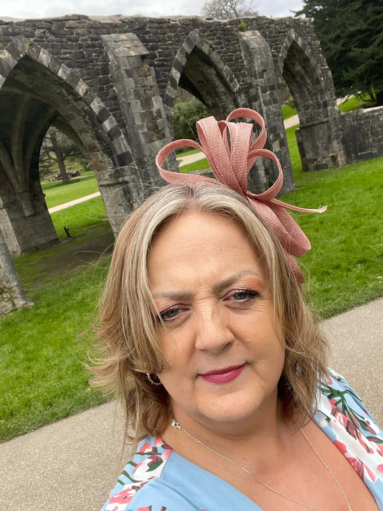 Sue Thomas in a pink fascinator, outside a ruined abbey.